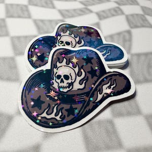 Skull cowboy hat sticker leaning against a stacked pile of matching stickers on a grey checkered background.