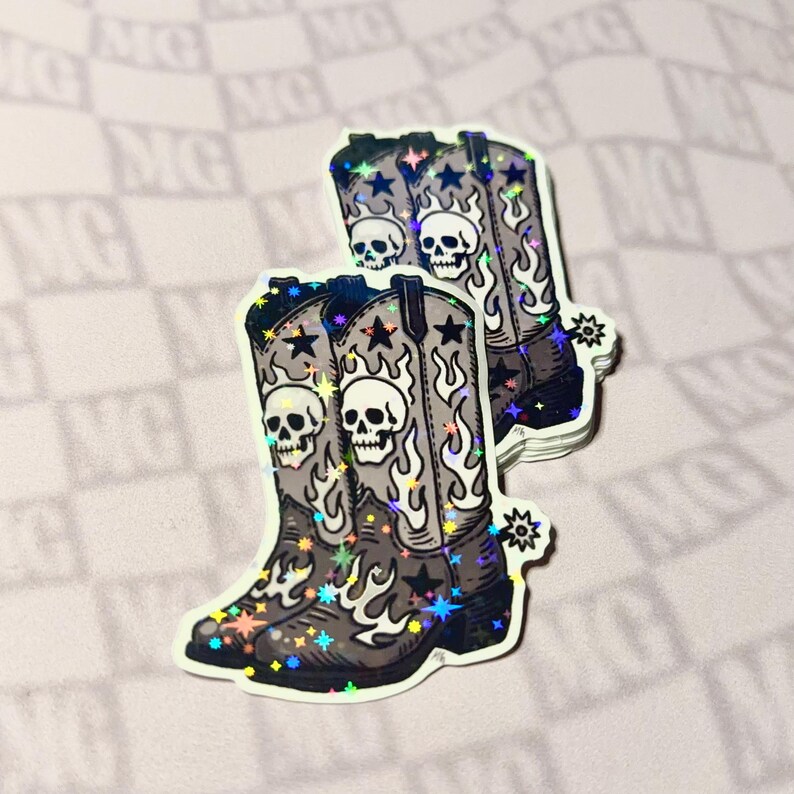 A grayscale vinyl sticker of cowboy boots with skulls, stars, and flames leaning against a small stack of identical stickers. The design features detailed Western motifs with a y’allternative, gothic country aesthetic.