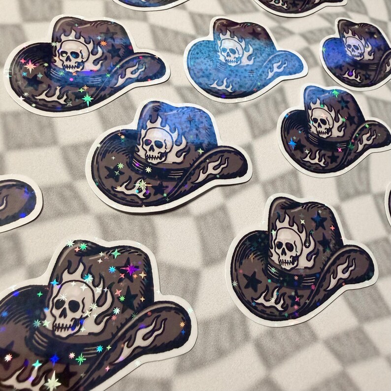 Multiple scattered holographic cowboy hat stickers with skull and flame designs on a grey checkered surface
