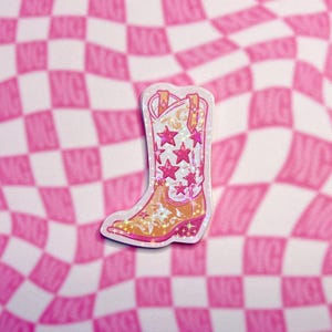 May include: A sticker of a cowboy boot with a pink and gold design. The boot features pink star accents and decorative floral patterns. The background is a pink and white checkered pattern.