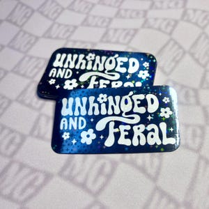 May include: Two rectangular stickers with the words "UNHINGED AND FERAL" in white, stylized font. The stickers have a dark blue background with a holographic effect, featuring white flowers and stars. The stickers are on a light pink surface.