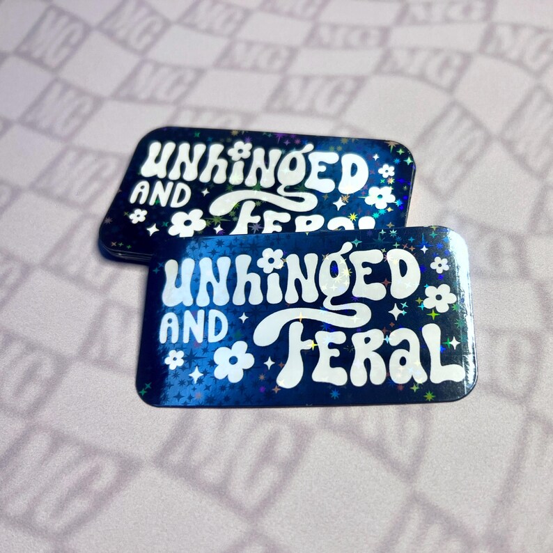 May include: Two rectangular stickers with the words "UNHINGED AND FERAL" in white, stylized font. The stickers have a dark blue background with a holographic effect, featuring white flowers and stars. The stickers are on a light pink surface.