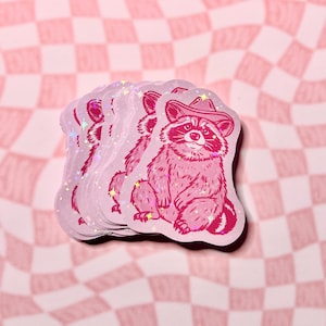 May include: A stack of pink and white holographic stickers featuring a cartoon raccoon wearing a cowboy hat. The raccoon is sitting down with its paws crossed and has a friendly expression.