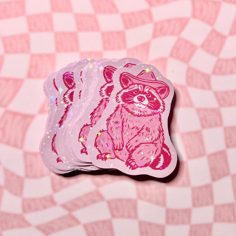 May include: A stack of pink and white holographic stickers featuring a cartoon raccoon wearing a cowboy hat. The raccoon is sitting down with its paws crossed and has a friendly expression.