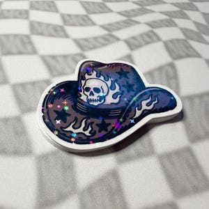 Close-up of an emo revival skull cowboy hat sticker with holographic sparkle on a greyscale checkered surface.