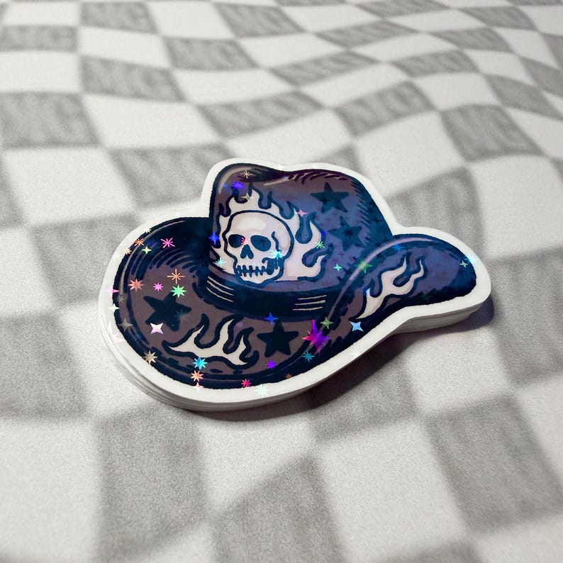 Close-up of an emo revival skull cowboy hat sticker with holographic sparkle on a greyscale checkered surface.