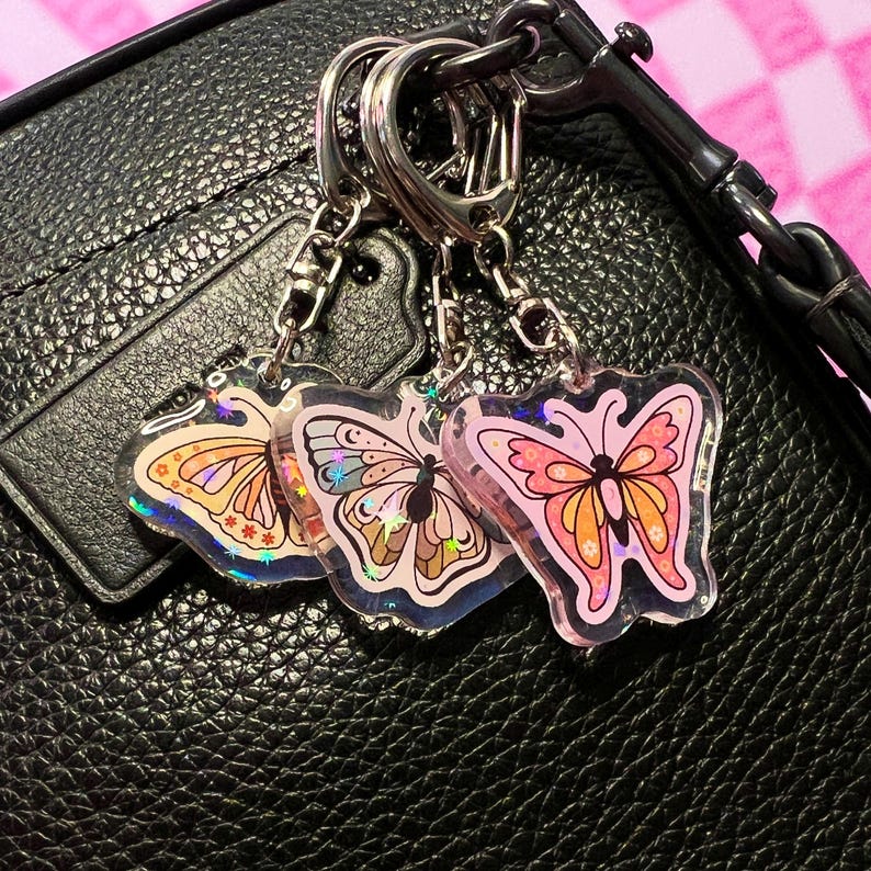 May include: Three iridescent butterfly keychains with pink, orange, and purple accents. The keychains are attached to a silver key ring.