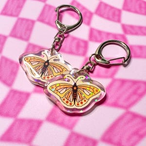 May include: Two clear keychains with a butterfly design. The butterflies are orange and yellow with black markings and a glittery background. The keychains are attached to silver key rings.