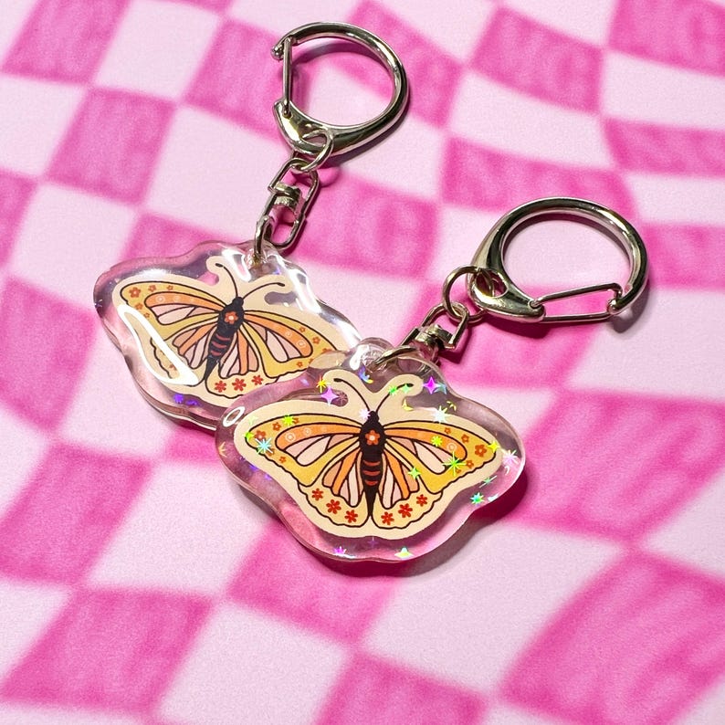 May include: Two clear keychains with a butterfly design. The butterflies are orange and yellow with black markings and a glittery background. The keychains are attached to silver key rings.