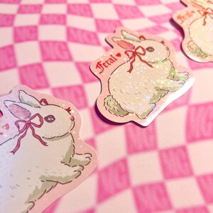 Peut inclure: Plusieurs autocollants repr&eacute;sentant un lapin blanc avec un n&oelig;ud rouge et le mot "Feral". Les autocollants ont un effet holographique et scintillant, sur un fond &agrave; carreaux roses et blancs. Id&eacute;al pour la d&eacute;coration.