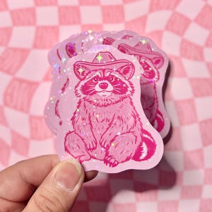 May include: Pink glitter sticker featuring a cartoon raccoon wearing a pink cowboy hat. The raccoon is sitting with its paws crossed and has a friendly expression.