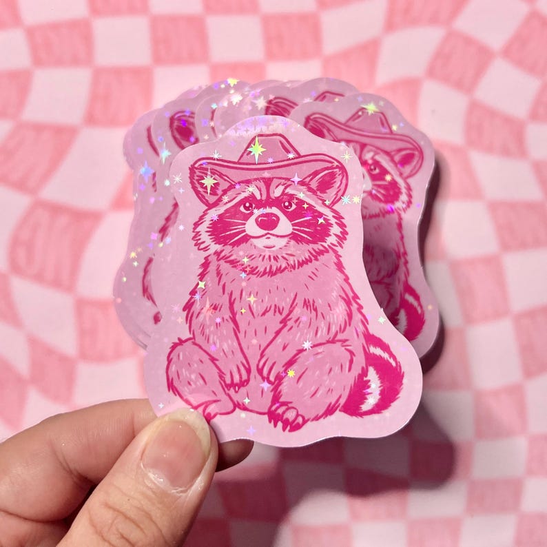 May include: Pink glitter sticker featuring a cartoon raccoon wearing a pink cowboy hat. The raccoon is sitting with its paws crossed and has a friendly expression.