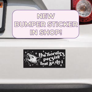 May include: A rectangular black bumper sticker featuring a white illustration of a screaming opossum surrounded by small stars. The text on the sticker reads, the horrors persist but so do i, written in a stylized white font against the black background.