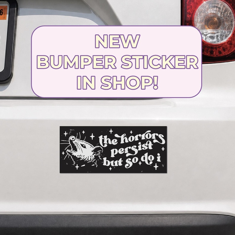 May include: A rectangular black bumper sticker featuring a white illustration of a screaming opossum surrounded by small stars. The text on the sticker reads, the horrors persist but so do i, written in a stylized white font against the black background.
