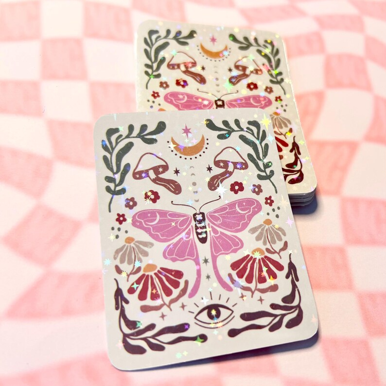 May include: A holographic sticker with a pink butterfly, mushrooms, flowers, and a crescent moon. The sticker has a white background and is surrounded by a green leafy border.