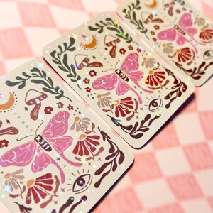 May include: Three holographic stickers with a whimsical design featuring pink butterflies, red and brown flowers, mushrooms, and stars on a white background.