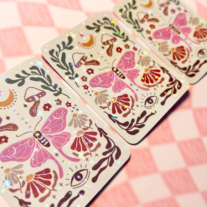 May include: Three holographic stickers with a whimsical design featuring pink butterflies, red and brown flowers, mushrooms, and stars on a white background.