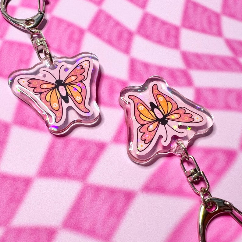 May include: Two pink butterfly keychains with glitter accents. Each keychain features a pink butterfly with orange wings and floral details. The keychains are attached to silver key rings.