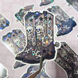 Close-up of a gothic Western cowboy boot sticker in black and gray tones, decorated with skulls, stars, and flames. The image captures a moody, alt-country aesthetic perfect for dark western lovers.