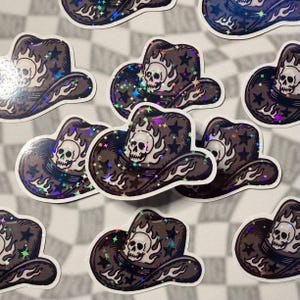 Multiple holographic sticker shaped like a cowboy hat with a skull in the center on a grey checkered background.
