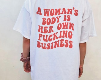 May include: A white short-sleeved t-shirt featuring red text on the back that reads A WOMAN'S BODY IS HER OWN FUCKING BUSINESS in a bold, wavy, retro-inspired font. The shirt has a loose, relaxed fit and a simple crew neckline.