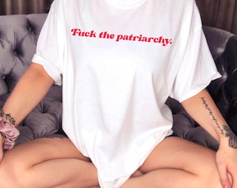 May include: A white short-sleeved t-shirt featuring the text "Fuck the patriarchy." printed in a red, cursive-style font across the chest. The garment has a crew neckline and a loose, relaxed fit with cuffed sleeves, made of a solid white fabric.