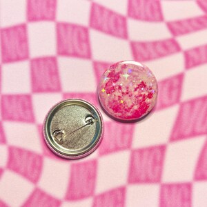 May include: Two round, pink and glittery button badges. One badge shows the front with a pink and white design and glitter. The other badge shows the back with a safety pin clasp. The background is a pink and white checkered pattern.