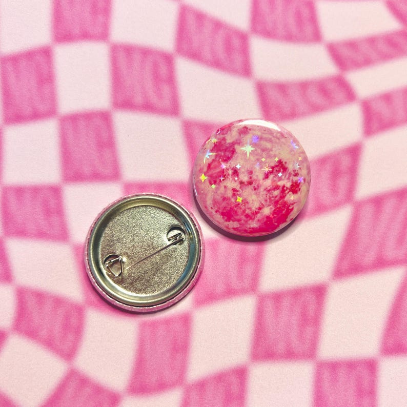May include: Two round, pink and glittery button badges. One badge shows the front with a pink and white design and glitter. The other badge shows the back with a safety pin clasp. The background is a pink and white checkered pattern.