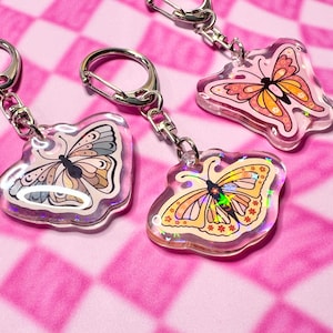 May include: Three butterfly keychains with clear acrylic charms. Each charm features a different butterfly design with pink, orange, and yellow colors. The butterflies have intricate details and floral patterns. The keychains are attached to silver metal rings.