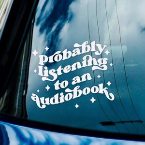 May include: A white vinyl sticker on a blue car window with the text "probably listening to an audiobook" in a retro font with stars around the text.