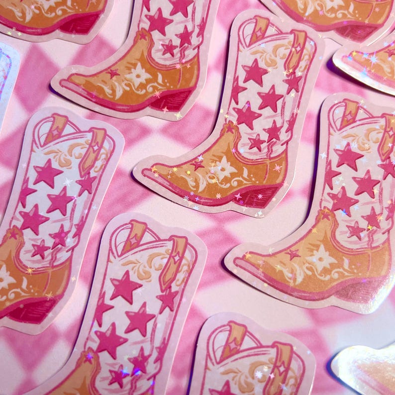May include: A collection of stickers featuring a cowboy boot design. The boots are primarily orange and white, with pink star and floral accents. The stickers have a holographic, glittery finish and are arranged on a pink and white checkered background.