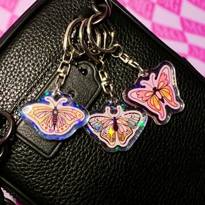 May include: Three iridescent butterfly keychains with gold chain links. The butterflies are pink, orange, and purple with glitter accents.