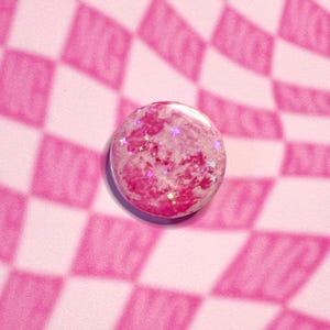 May include: A circular, pink button with a marbled design and shimmering star-shaped glitter. The button is placed on a pink and white chequered background, giving a fun and quirky look.