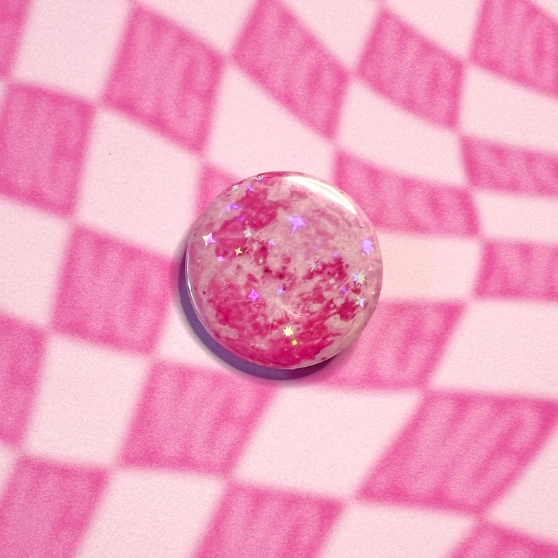 May include: A circular, pink button with a marbled design and shimmering star-shaped glitter. The button is placed on a pink and white chequered background, giving a fun and quirky look.