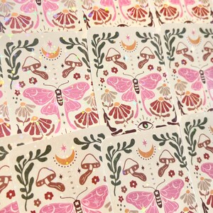 May include: A set of holographic stickers featuring a pink butterfly, mushrooms, flowers, and a crescent moon on a cream background.