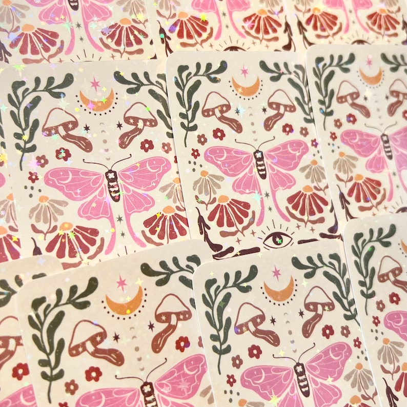 May include: A set of holographic stickers featuring a pink butterfly, mushrooms, flowers, and a crescent moon on a cream background.