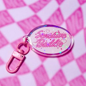 May include: A pink keychain with the words "Sensitive Baddie" in a cursive font, decorated with hearts and stars. The keychain has a pink clasp and a clear, oval-shaped charm with a holographic effect, set against a pink checkered background.