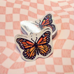 May include: Two iridescent holographic stickers of a butterfly with orange and black wings. The butterfly has a black body and a white background. The stickers are on a pink and white checkered background.