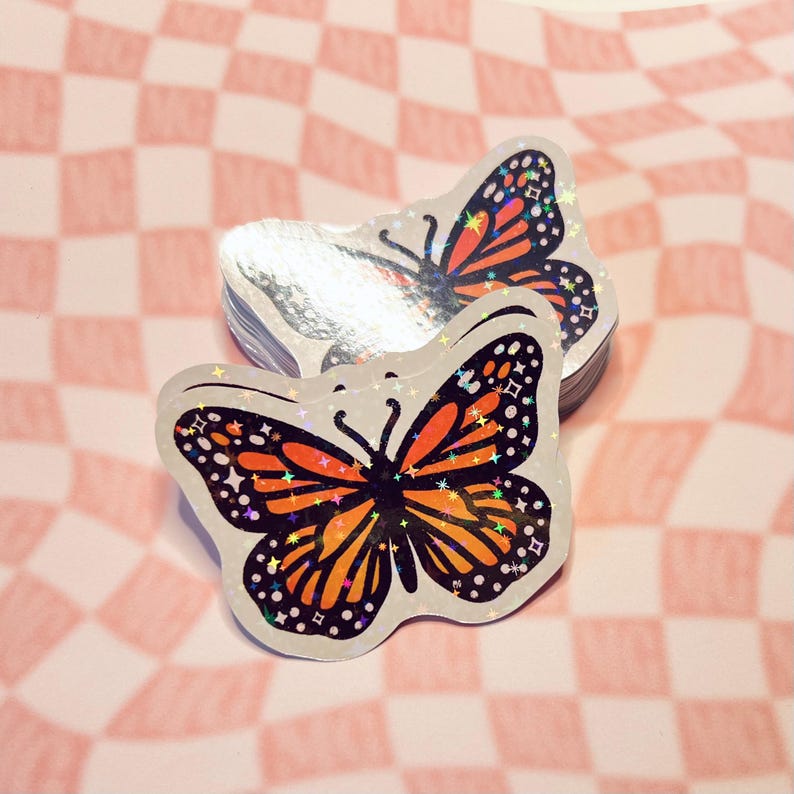 May include: Two iridescent holographic stickers of a butterfly with orange and black wings. The butterfly has a black body and a white background. The stickers are on a pink and white checkered background.