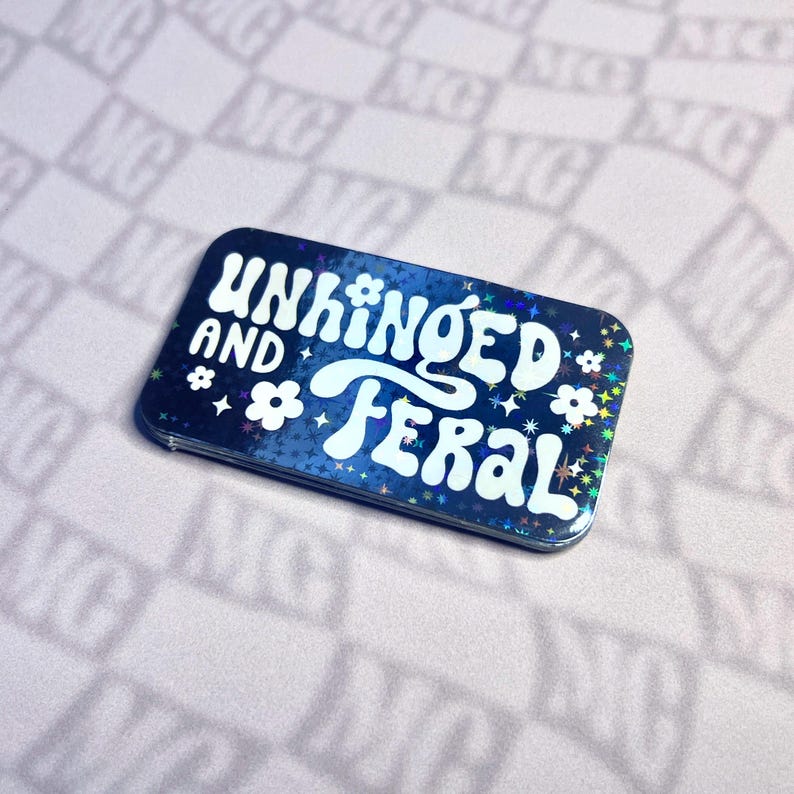 May include: A rectangular sticker with rounded corners, featuring the words "UNHINGED AND FERAL" in white, stylized font. The sticker has a dark blue background with a holographic effect, adorned with white stars and flower graphics.