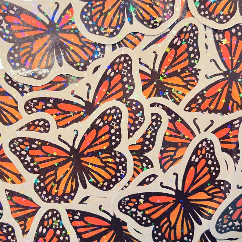 May include: A close-up of a pile of holographic butterfly stickers. The butterflies are orange, black, and iridescent. The stickers are on a light brown background.