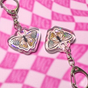 May include: Two iridescent butterfly keychains with a silver key ring. The butterflies are a light pink, blue, and yellow with a white background. The keychains are on a pink and white checkered background.