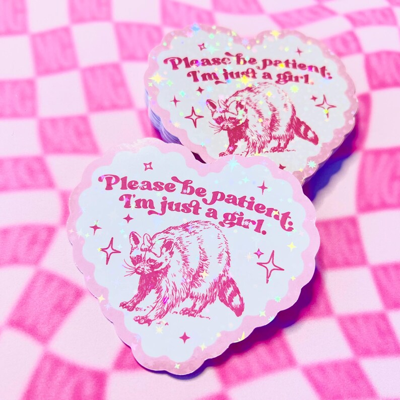 Peut inclure: Un autocollant en forme de c&oelig;ur rose avec une illustration de raton laveur et le texte "Please be patient, I'm just a girl." L'autocollant a une finition paillet&eacute;e et se trouve sur un fond &agrave; carreaux rose et blanc.