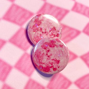 May include: Two round, pink resin buttons with a marbled effect and embedded glitter. The buttons are a vibrant pink colour with white and darker pink swirls, and sit on a pink and white chequered background.