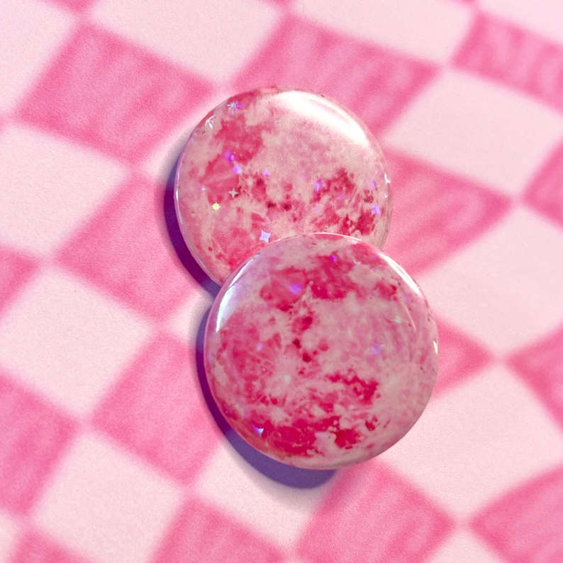 May include: Two round, pink resin buttons with a marbled effect and embedded glitter. The buttons are a vibrant pink colour with white and darker pink swirls, and sit on a pink and white chequered background.