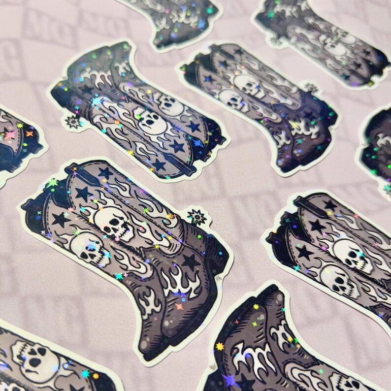Multiple grayscale cowboy boot stickers scattered on a neutral gray background. The skull and flame designs catch the light, showing their Western-gothic style and alt-country inspiration.