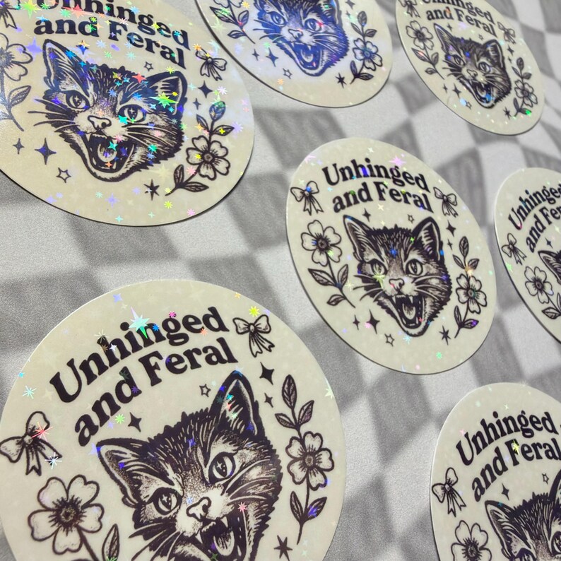May include: A collection of round stickers with a cat illustration and the text "Unhinged and Feral". The cat is depicted with an open mouth and sharp teeth, surrounded by floral and star designs. The stickers have a holographic, iridescent finish.
