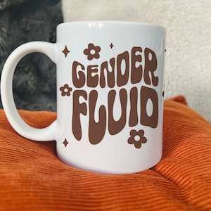 May include: White ceramic mug with a large handle, featuring the words "GENDER FLUID" in brown, retro-style lettering. The design includes small flower and diamond shapes. The mug is set against an orange cushion and a gray, textured background.