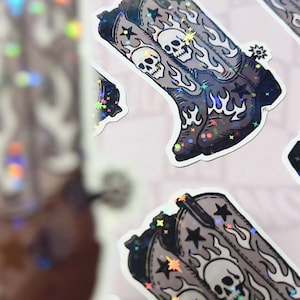 A grayscale vinyl sticker with skull-covered cowboy boots surrounded by stars and flames, photographed on a dark gray background. The composition evokes a Southern gothic mood with vintage Western edge.