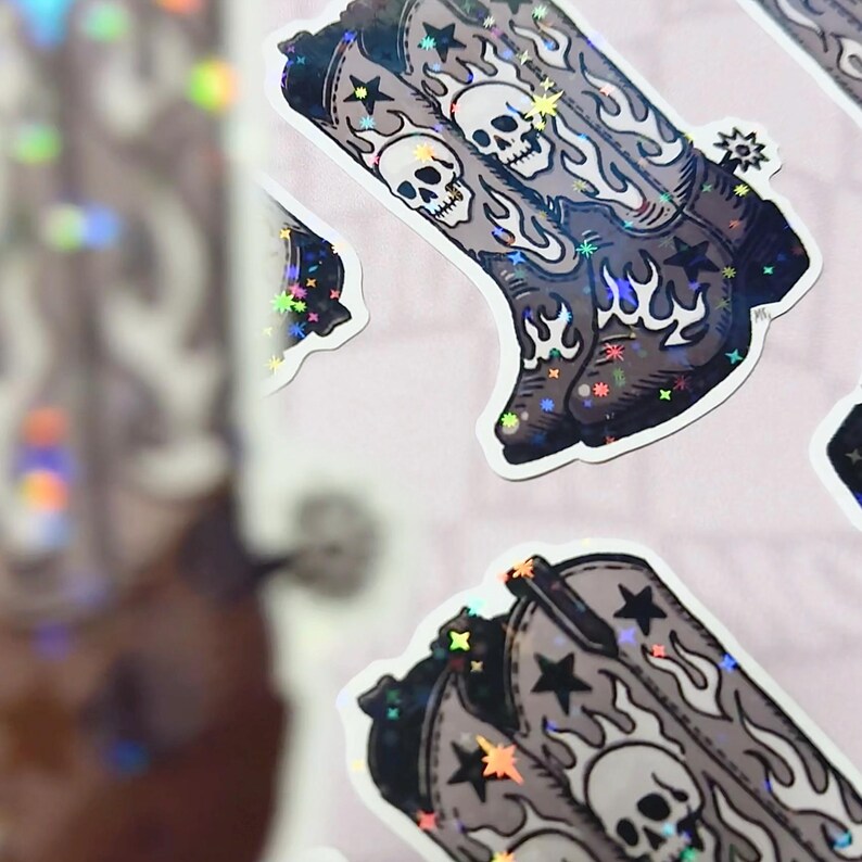 A grayscale vinyl sticker with skull-covered cowboy boots surrounded by stars and flames, photographed on a dark gray background. The composition evokes a Southern gothic mood with vintage Western edge.